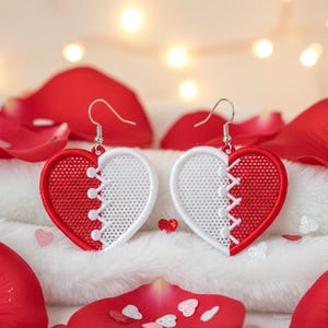 Puede incluir: Pendientes en forma de corazón, divididos en mitades rojas y blancas, con un diseño cosido. Los pendientes cuelgan de ganchos plateados, sobre un fondo de pétalos de rosa rojos y tela blanca suave.