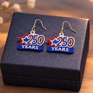 May include: A pair of patriotic earrings with gold-colored hooks. Each earring features a red, white, and blue star design with the number "250" and the word "YEARS" on a blue banner. The earrings are displayed on a black jewelry box.