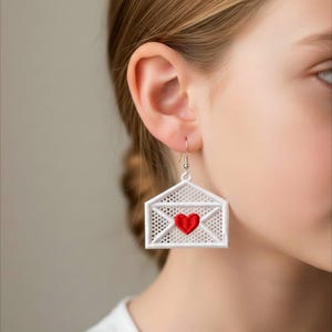 May include: White embroidered envelope earrings with a red heart in the center. The earrings are hanging from silver-colored ear wires. The earrings are a unique and romantic accessory.
