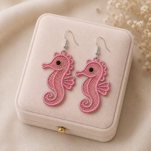 May include: A pair of dangle earrings featuring pink seahorse-shaped lace motifs. Each seahorse has a small black bead for an eye and is attached to a silver-toned metal fishhook ear wire. The lace design displays a fine, intricate mesh pattern.