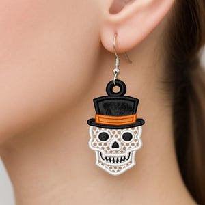 May include: A single Halloween earring featuring a white skull with a black top hat and orange band. The skull has embroidered details, including eye sockets, a nose, and teeth. The earring is suspended from a silver hook.