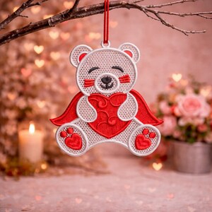 May include: Embroidered teddy bear ornament with a red heart and cape. The bear is white with red accents, including a heart-shaped chest and paw prints. It hangs from a red string, perfect for Valentine's Day or a special occasion.