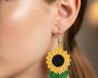 Puede incluir: Pendientes de girasol bordados con pétalos amarillos, centro negro y hojas verdes. Los pendientes cuelgan de ganchos plateados. El diseño es un accesorio floral detallado.