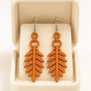 May include: A pair of orange leaf-shaped earrings with silver hooks, presented in a white jewellery box. The earrings feature a detailed, textured design, resembling a fern. The earrings are a warm, earthy tone.