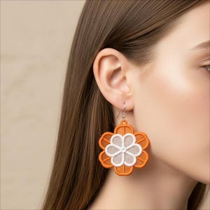 May include: Orange and white flower-shaped earrings with a detailed embroidered design. The earrings are hanging from silver hooks. The flower has a layered design with a white center and orange outer petals.