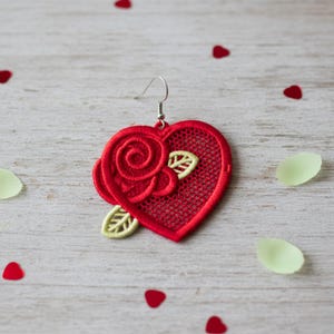 May include: A red heart-shaped earring with a rose and leaf design. The earring features a delicate, openwork pattern and a silver-colored hook. Scattered around the earring are red heart-shaped confetti and green petal-shaped confetti.