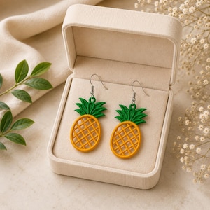 Può includere: Un paio di orecchini pendenti a forma di ananas con un corpo giallo a motivo incrociato e una parte superiore a foglie verdi. Gli orecchini sono sospesi a ganci in metallo color argento e sono esposti in un portagioie rettangolare color crema.