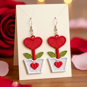以下が含まれることがあります： 刺繍されたハート型のイヤリング。各イヤリングは、赤いハート型の花と緑の葉が赤いハートの白いポットに入っています。イヤリングは銀色のフックから吊り下げられています。イヤリングはクリーム色のカードに表示されています。
