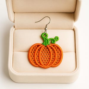 May include: An orange pumpkin-shaped earring with green stem and tendrils. The earring is made of embroidered thread and has a silver-colored hook. The earring is displayed inside a beige jewelry box.