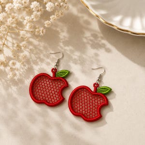 May include: A pair of dangle earrings shaped like red apples with a honeycomb-patterned cutout center and a small green leaf at the top. The earrings feature silver-toned metal hooks for attachment and are crafted with detailed red embroidery.