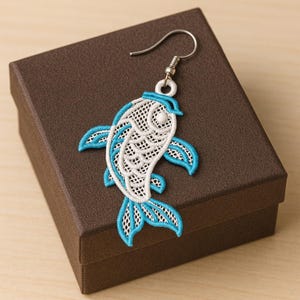 Puede incluir: Un solo pendiente con forma de pez con un gancho plateado. El pendiente presenta un diseño blanco de tejido abierto con detalles en azul en las aletas y la cabeza. El pendiente se muestra en una caja marrón.