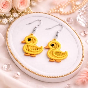 May include: A pair of yellow duck earrings with black eyes and orange beaks. The earrings are made of a textured material and have silver-colored hooks. The earrings are displayed on a white plate with a gold rim.