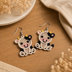 May include: A pair of dangle earrings featuring embroidered cow charms. Each charm is crafted with a cream-colored mesh body, black patches, a pink snout, and small yellow horns. The cow figures are attached to silver-toned metal earring hooks.