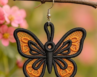 May include: A single butterfly earring with a black outline and orange filigree wings hangs from a silver hook. The earring is suspended from a brown branch against a blurred background of pink flowers and green foliage.