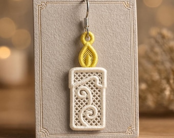 Puede incluir: Un solo pendiente de vela con una llama amarilla y un cuerpo de vela bordado en blanco. El pendiente está sujeto a un gancho plateado y se muestra en una tarjeta gris con bordes decorativos. El diseño de la vela presenta detalles intrincados.