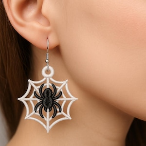 Puede incluir: Un par de pendientes con temática de Halloween. Cada pendiente presenta un diseño de telaraña blanca con una araña negra en el centro. Los pendientes están suspendidos de un gancho plateado.