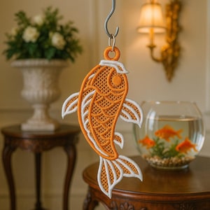 Könnte beinhalten: Ein orange-weiß bestickter Koi-Fisch-Ornament, der an einem silbernen Haken hängt. Der Fisch hat detaillierte Schuppenmuster und weiße Akzente auf seinen Flossen und Schwanz. Im Hintergrund ein Fischglas mit Goldfischen und eine dekorative Vase.