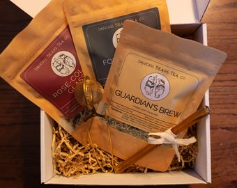 May include: A gift box containing three tea pouches with the Dancing Bears Tea Co. logo. The pouches are labeled "Rose-Col Glass", "Fo", and "Guardian's Brew". A gold tea infuser and cinnamon stick are also included.