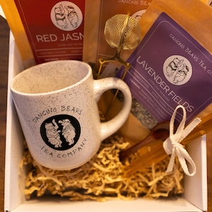 Może przedstawiać: Pudełko upominkowe zawierające nakrapiany biały kubek z logo Dancing Bears Tea Company, torebki herbaty, zaparzacz do herbaty i patyczki miodu. Torebki herbaty są oznaczone jako "Red Jasmine" i "Lavender Fields". Pudełko jest wypełnione pociętym papierem.