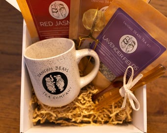 以下が含まれることがあります： Dancing Bears Tea Companyのロゴが入った、白い斑点のあるマグカップ、ティーバッグ、ティーインフューザー、ハニースティックが入ったギフトボックス。 ティーバッグには「Red Jasmine」と「Lavender Fields」と書かれています。 箱は細かく刻んだ紙で満たされています。