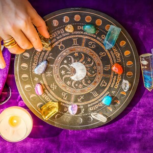 Peut inclure: Une roue astrologique marron fonc&eacute; avec des symboles du zodiaque et un croissant de lune, entour&eacute;e de cristaux color&eacute;s et d'une bougie allum&eacute;e. Une main avec des bagues dor&eacute;es place un cristal sur la roue. La roue est sur une surface de velours violet.
