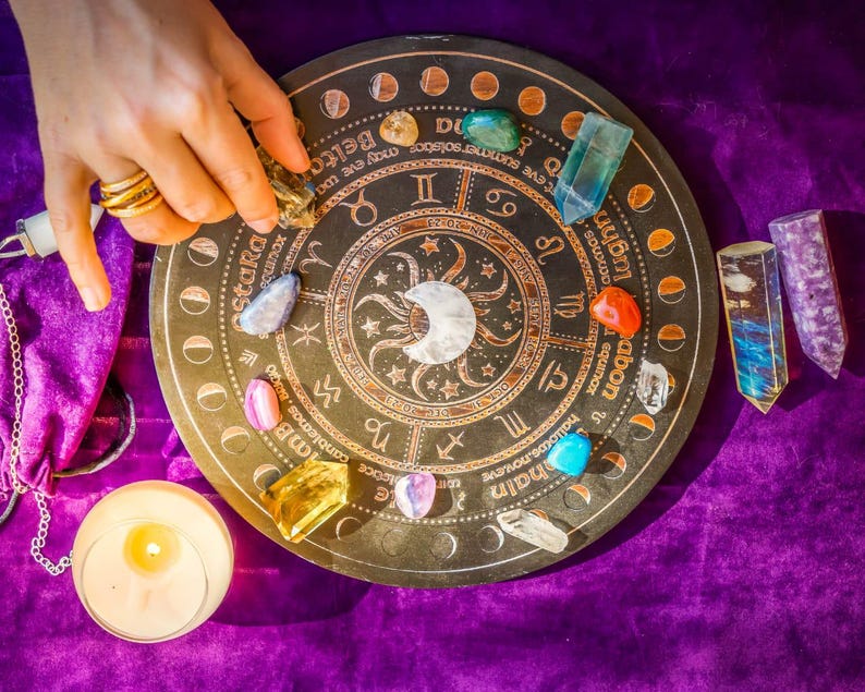Peut inclure: Une roue astrologique marron fonc&eacute; avec des symboles du zodiaque et un croissant de lune, entour&eacute;e de cristaux color&eacute;s et d'une bougie allum&eacute;e. Une main avec des bagues dor&eacute;es place un cristal sur la roue. La roue est sur une surface de velours violet.