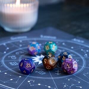 Peut inclure: Plusieurs d&eacute;s poly&eacute;driques color&eacute;s avec des chiffres dor&eacute;s, pos&eacute;s sur un tableau astrologique bleu fonc&eacute;. Les d&eacute;s sont de diff&eacute;rentes couleurs, notamment bleu, violet, vert et marron. Une bougie allum&eacute;e dans un verre est en arri&egrave;re-plan.