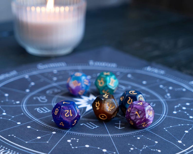 Peut inclure: Plusieurs d&eacute;s poly&eacute;driques color&eacute;s avec des chiffres dor&eacute;s, pos&eacute;s sur un tableau astrologique bleu fonc&eacute;. Les d&eacute;s sont de diff&eacute;rentes couleurs, notamment bleu, violet, vert et marron. Une bougie allum&eacute;e dans un verre est en arri&egrave;re-plan.