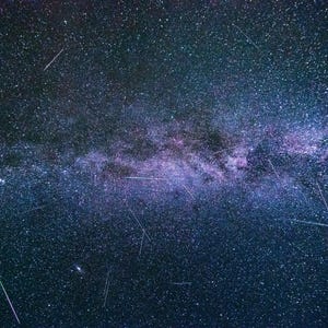 May include: A night sky photograph showcasing the Milky Way galaxy with numerous stars and streaks of light. The image features a dark blue and purple color palette, with the Milky Way appearing as a bright, central band.