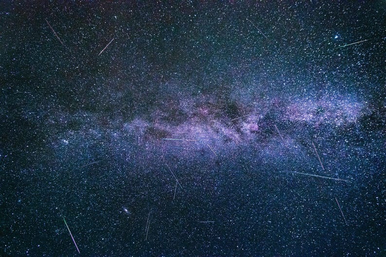 May include: A night sky photograph showcasing the Milky Way galaxy with numerous stars and streaks of light. The image features a dark blue and purple color palette, with the Milky Way appearing as a bright, central band.