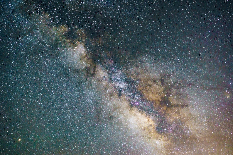 May include: A photograph of the night sky, showcasing the Milky Way galaxy. The image captures a vast expanse of space filled with countless stars, with a prominent band of light and color. The colors range from deep blues and greens to warm yellows and browns.