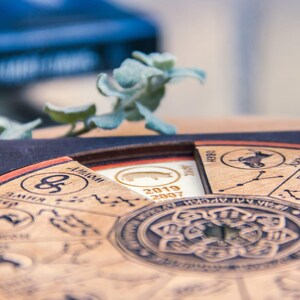 Peut inclure: Calendrier du zodiaque en bois avec des symboles astrologiques et du texte grav&eacute;s. Le calendrier pr&eacute;sente un design circulaire avec un motif floral central et une section amovible avec les ann&eacute;es 2019 et 2007. Une petite plante succulente est en arri&egrave;re-plan.