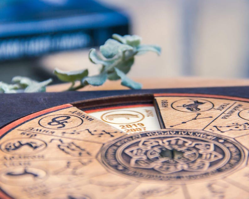 Peut inclure: Calendrier du zodiaque en bois avec des symboles astrologiques et du texte grav&eacute;s. Le calendrier pr&eacute;sente un design circulaire avec un motif floral central et une section amovible avec les ann&eacute;es 2019 et 2007. Une petite plante succulente est en arri&egrave;re-plan.