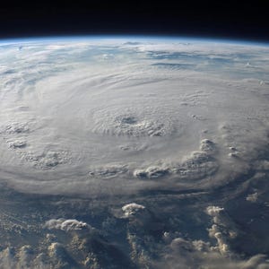 May include: An aerial view of a large hurricane swirling over the Earth. The storm's eye is visible at the center, surrounded by swirling cloud formations. The image shows the curvature of the Earth and the dark expanse of space.