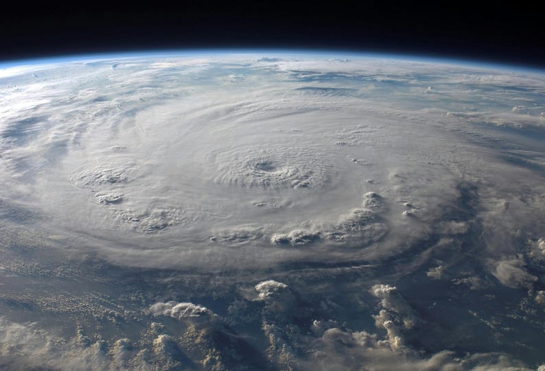 May include: An aerial view of a large hurricane swirling over the Earth. The storm's eye is visible at the center, surrounded by swirling cloud formations. The image shows the curvature of the Earth and the dark expanse of space.