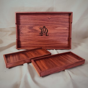 May include: A set of three wooden trays in varying sizes. The large tray features a carved monogram. The wood has a rich reddish-brown color and a visible grain pattern. The trays are rectangular with raised edges.