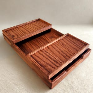 May include: A tiered wooden tray set with a rich, reddish-brown stain. The set includes a small, rectangular tray on top, a larger tray in the middle, and a bottom tray with a pull-out drawer. The wood grain is visible.