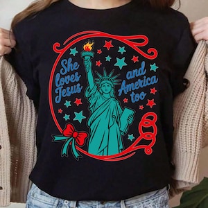 Peut inclure: T-shirt noir avec un motif de la Statue de la Liberté turquoise tenant une torche et une tablette, entourée d'étoiles rouges et turquoise. Le texte dit "She loves Jesus and America too" en turquoise et rouge.