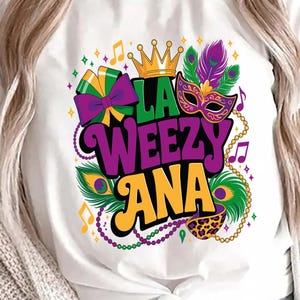 Peut inclure: T-shirt blanc avec un motif coloré de Mardi Gras. Le graphisme comprend le texte "LA WEEZY ANA" en violet et orange, une couronne dorée, un masque, des plumes, des notes de musique et un collier de perles.
