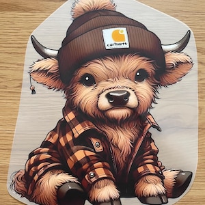 Peut inclure: Autocollant représentant une vache des Highlands de dessin animé portant un bonnet Carhartt marron avec un pompon et une veste à carreaux. La vache a de grands yeux et de petites cornes. Le logo Carhartt est visible sur le bonnet.