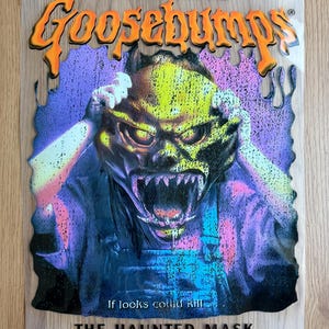 May include: A clear plastic Goosebumps book cover. It features a monster with a yellow and green mask, hands on its head, and a menacing expression. The title "Goosebumps" is in orange, and the book title is "The Haunted Mask."