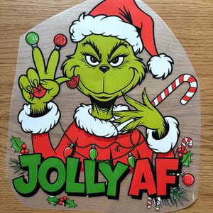 May include: A green cartoon character in a Santa hat and red coat, holding a candy cane and making a peace sign. The text "JOLLY AF" is displayed in green and red, with holly accents. Christmas-themed.