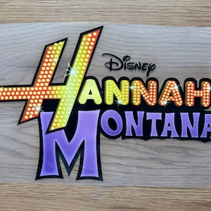 May include: A colourful Hannah Montana logo with the Disney logo above. The logo features the name "Hannah Montana" in yellow, orange, and red, with a purple "M" and black outlines. The design has a glittery effect.