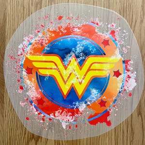 May include: A circular, transparent sticker displaying the Wonder Woman emblem in yellow, set against a blue and orange backdrop. Red star accents and paint splatter effects surround the logo, creating a dynamic and colourful design.