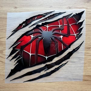 May include: A red and black Spider-Man graphic on a clear background. The design features a spider web pattern and a black spider silhouette. The image has a torn effect with black claw marks.