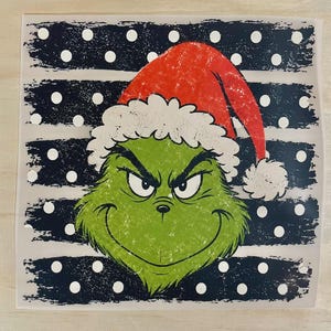 Peut inclure: Un autocollant carré représentant un personnage vert et poilu à l'expression renfrognée portant un bonnet de Père Noël rouge et blanc. Le fond se compose de rayures horizontales bleu foncé usées avec un motif répété de pois blancs.