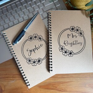 May include: Two spiral-bound notebooks with kraft paper covers. One notebook has the name "Sophie" inside a floral wreath, and the other says "Mrs. Reightley." A silver and black pen rests on one notebook.