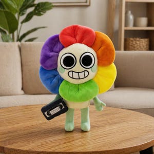 Könnte beinhalten: Ein Plüschtier in Form einer lächelnden Blume mit einem Regenbogen-Blütenblatt-Design. Die Blume hat große schwarze Augen, einen grünen Stiel und hält eine schwarze Kassette. Das Spielzeug steht auf einem Holztisch.