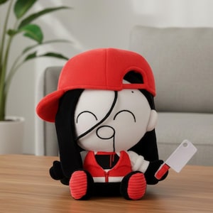 Puede incluir: Muñeco de peluche con gorra de béisbol roja, pelo negro y chaleco rojo. El muñeco tiene una cara blanca con un parche en el ojo negro y sostiene una pequeña cuchilla. El muñeco está sentado sobre una superficie de madera.