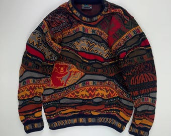 May include: A colorful, patterned sweater with a crew neck. The sweater features a mix of geometric and abstract designs in shades of red, orange, brown, gray, and navy blue. The text "BACK" and "KA DOU" are visible on the sweater.